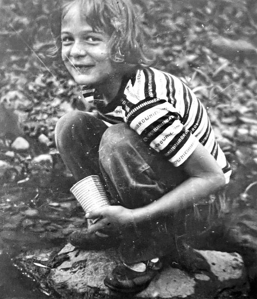Susan Robbins as a child, squatting at the side of a river and smiling at the camera