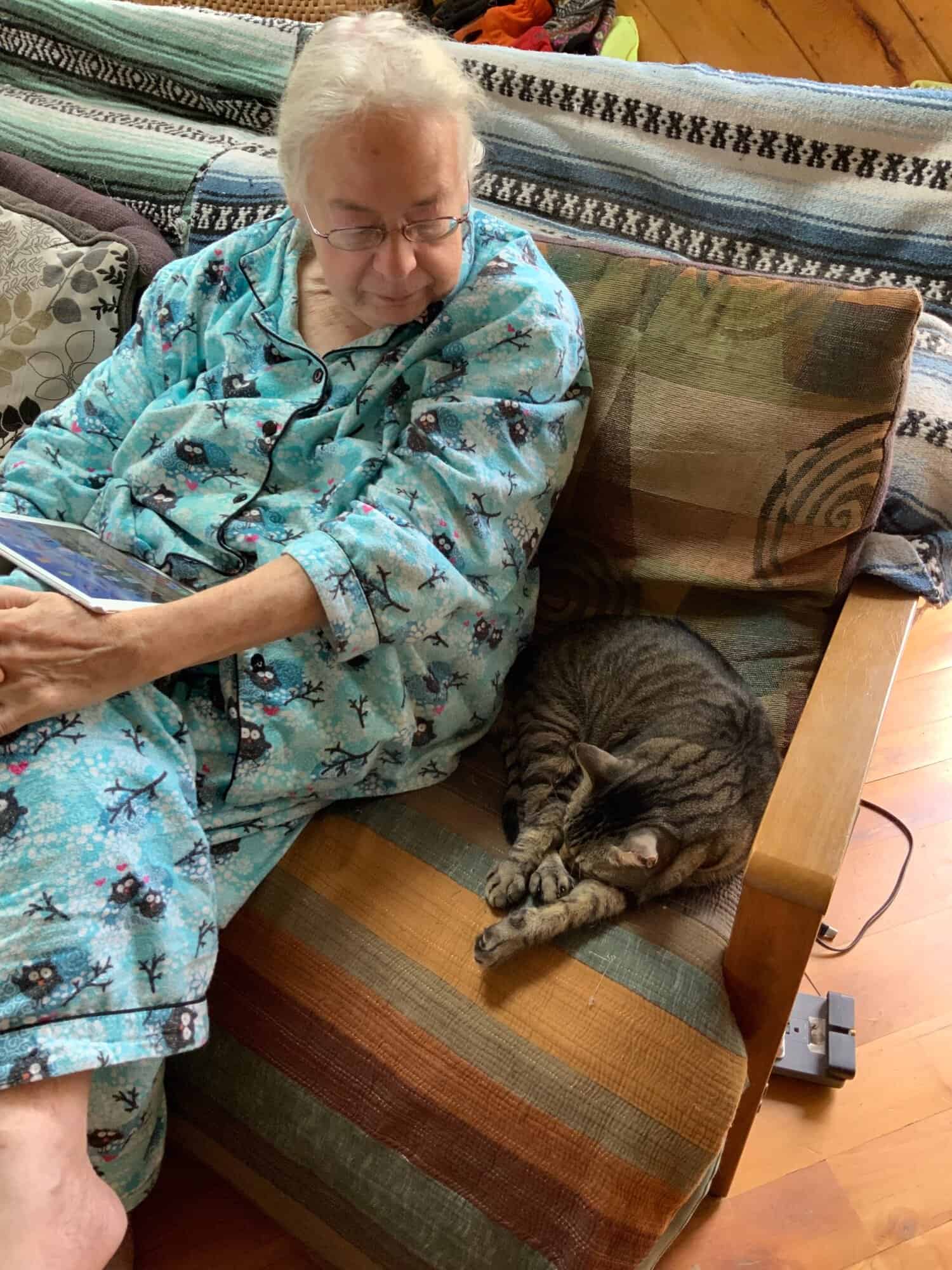 Susan sitting on a couch in pajamas looking at a gray tabby cat sitting beside her